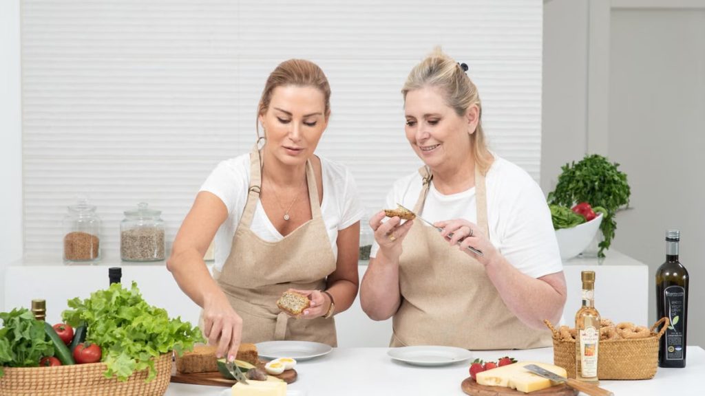 Two women prepare sandwiches with fresh vegetables and bread, enjoying a cooking session together indoors.